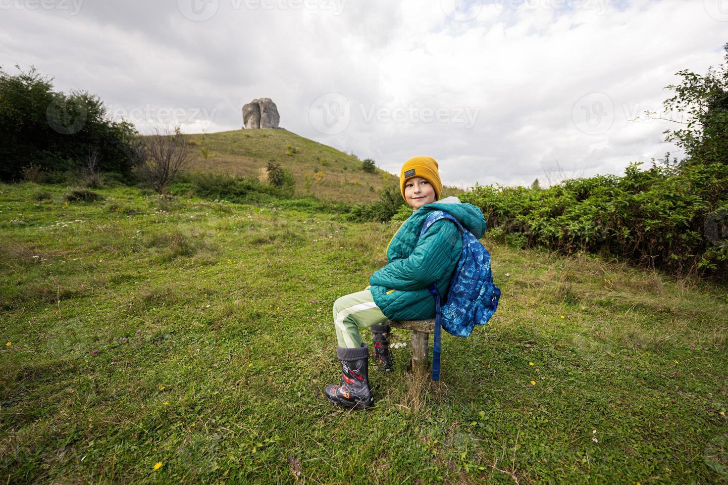 atividade no dia ensolarado de outono, menino explorando a natureza. garoto usa chapéu amarelo e mochila sente-se contra a grande pedra de pidkamin, ucrânia. foto