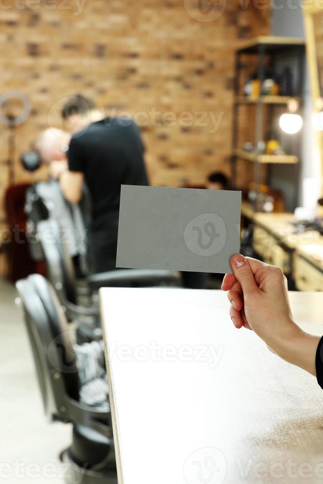 mão feminina segurando cartão postal cinza ou cartão de mandona com mock up no elegante fundo de barbearia ou salão de beleza. homem barbeiro turva está cortando e raspando a barba o cliente. foto