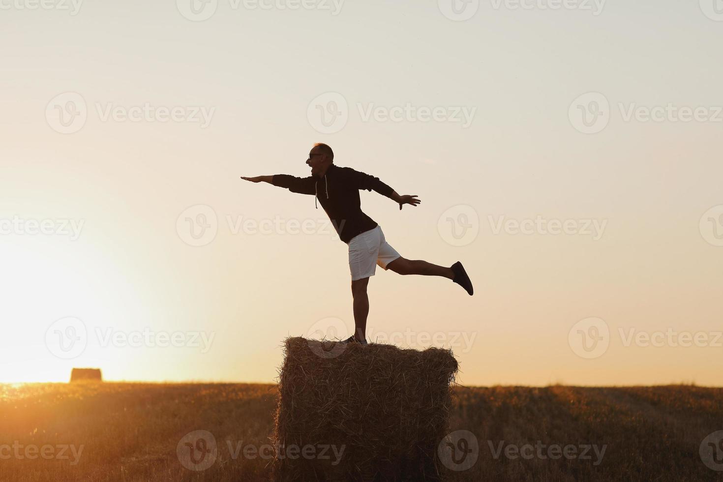 jovem bonito de moletom preto e short branco, no campo de verão ao ar livre, se divertindo no fardo de feno, palheiro no pôr do sol foto