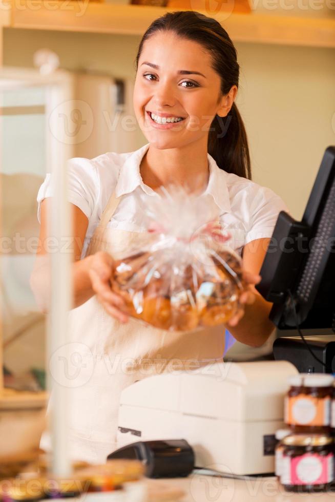 desfrute de biscoitos frescos linda jovem caixa feminina no avental em pé perto da caixa registradora e esticando os biscoitos foto