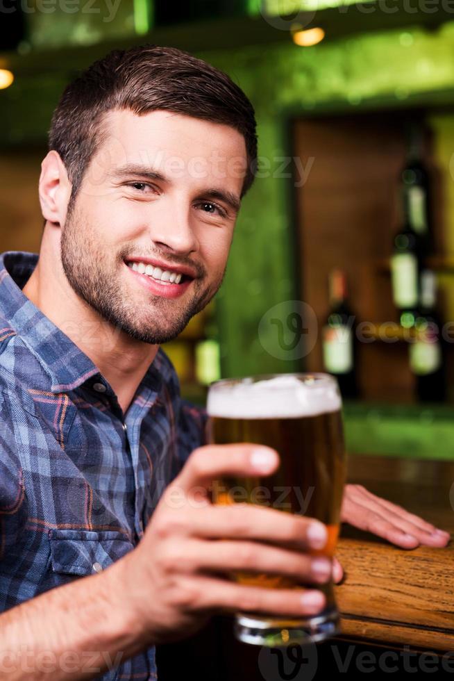 felicidades jovem bonito brindando com cerveja e olhando para a câmera com sorriso enquanto está sentado no balcão do bar foto