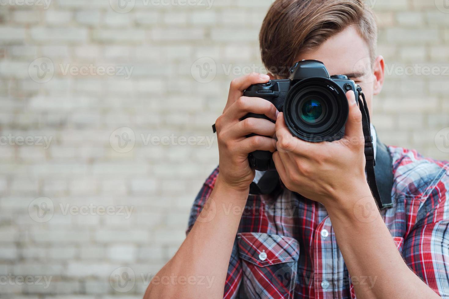 sorria para a câmera jovem confiante focando em você com sua câmera digital em pé contra a parede de tijolos foto