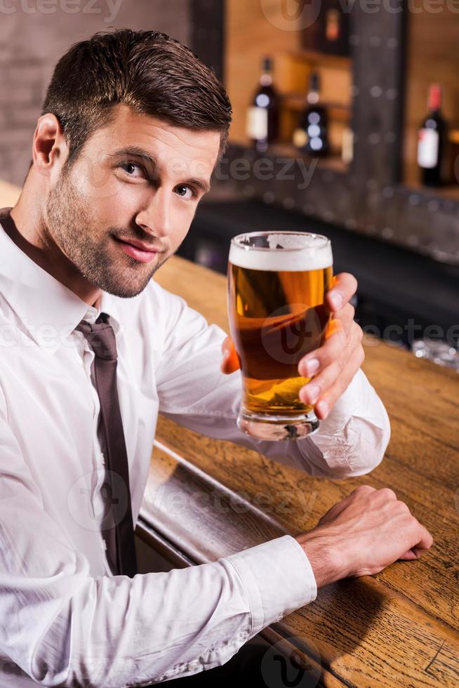 felicidades jovem bonito de camisa e gravata brindando com cerveja e olhando para a câmera enquanto está sentado no balcão do bar foto