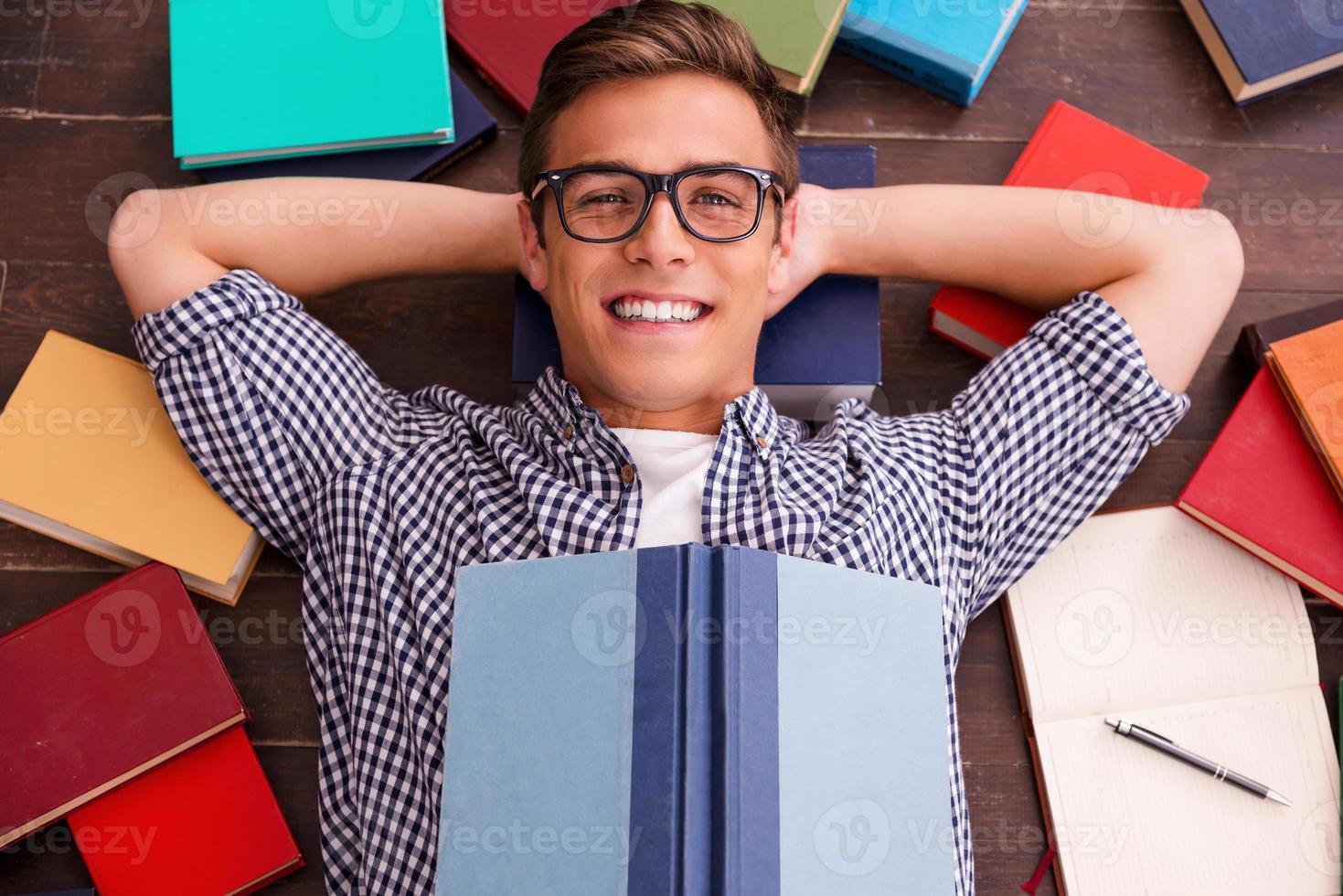 a leitura é o meu hobby vista superior do jovem feliz segurando as mãos atrás da cabeça e sorrindo enquanto estava deitado no chão de madeira com livros coloridos ao seu redor foto