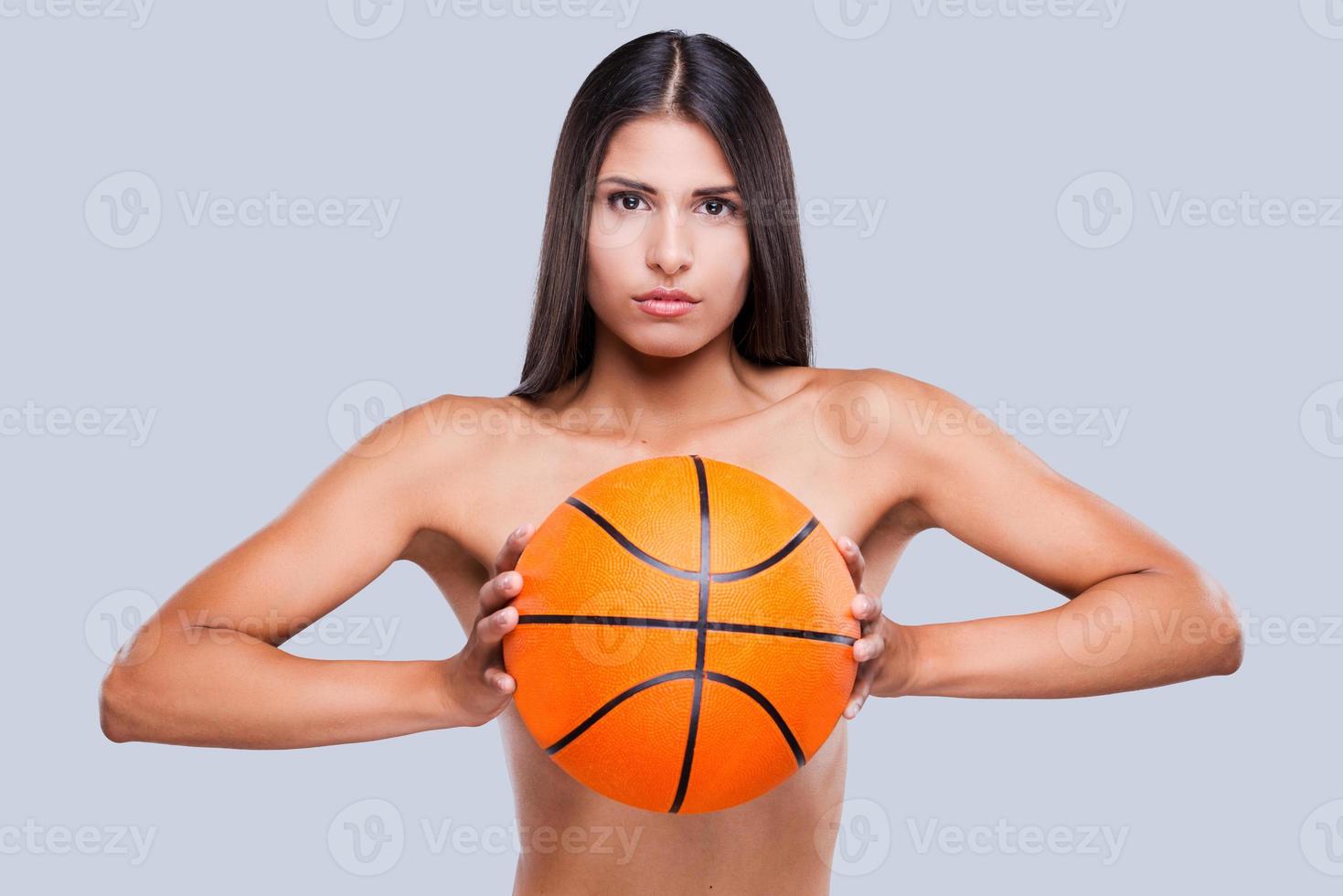 pegar a bola linda mulher sem camisa segurando uma bola de basquete e olhando para a câmera em pé contra um fundo cinza foto