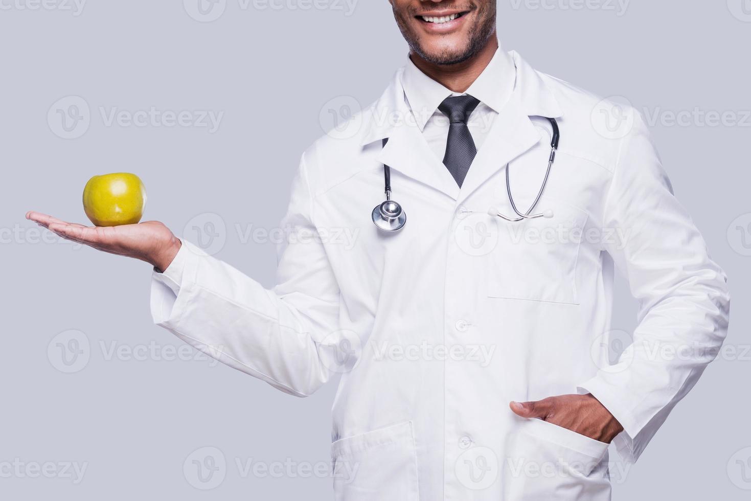 comer a imagem recortada saudável do médico africano esticando a maçã verde em pé contra um fundo cinza foto