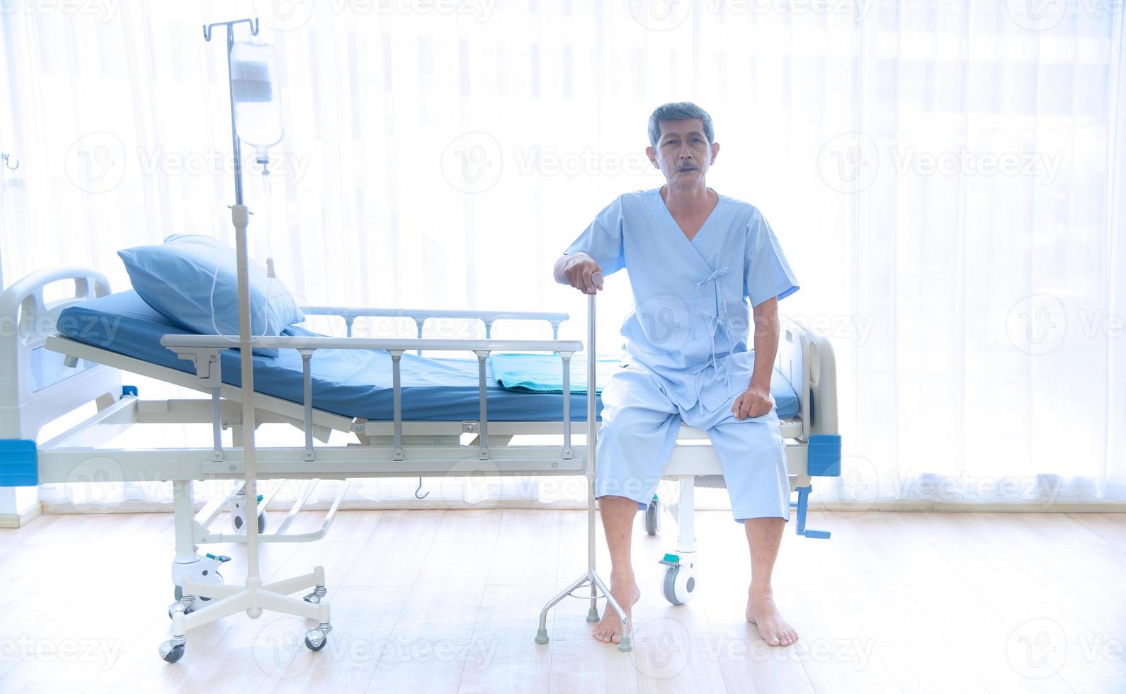 asiático doente ou idoso não podia andar sentado sozinho na cama do paciente com camisa azul e bengala esperando o médico e a enfermeira para tratamento ou cura no hospital ou assistência médica foto