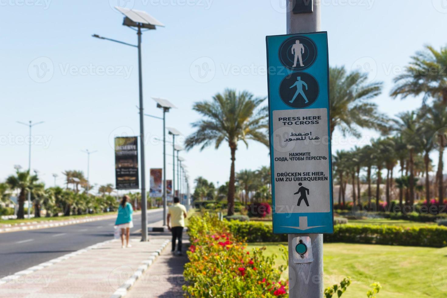 botão para chamar a passagem de pedestres com a seta para cima. clique aqui para cruzá-lo escrito em quatro idiomas inglês, árabe, russo e alemão foto