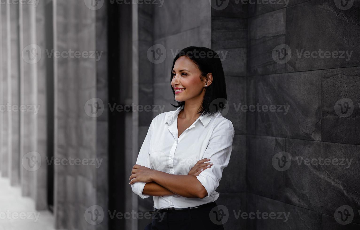 retrato de uma mulher de negócios de raças mistas sorridente, braço cruzado e desviar o olhar na cidade urbana foto