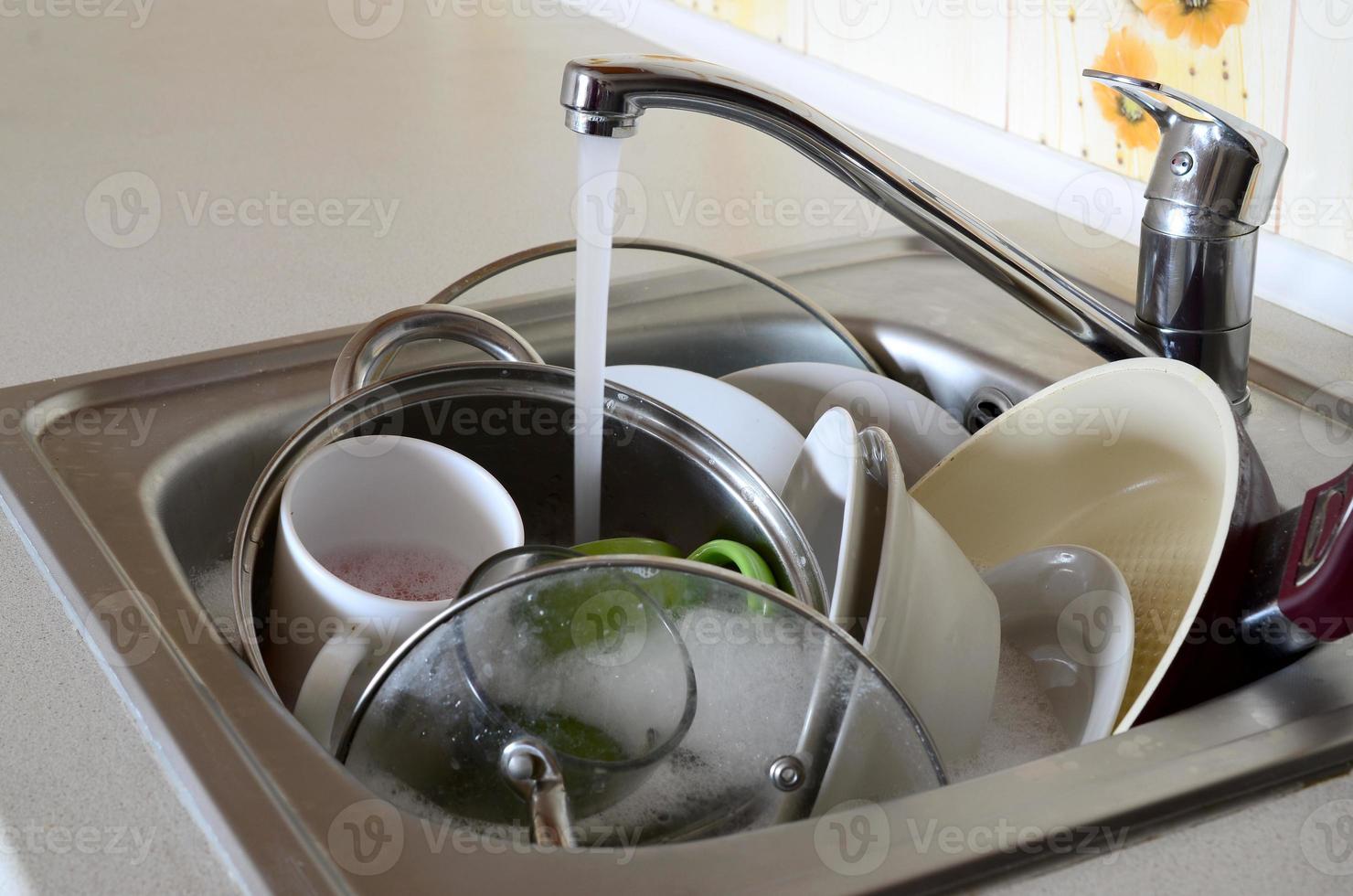 pratos sujos e utensílios de cozinha não lavados estão em água de espuma sob uma torneira de uma torneira de cozinha foto