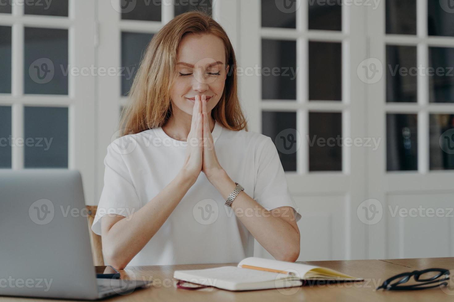 mulher sentada no laptop com as palmas das mãos entrelaçadas, meditando, controlando o estado emocional, fazendo uma pausa no trabalho foto