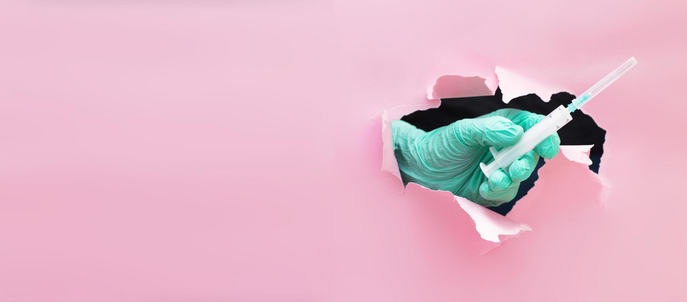 a mão do médico estende uma seringa com uma vacina através de um buraco em papel rasgado em um fundo rosa foto