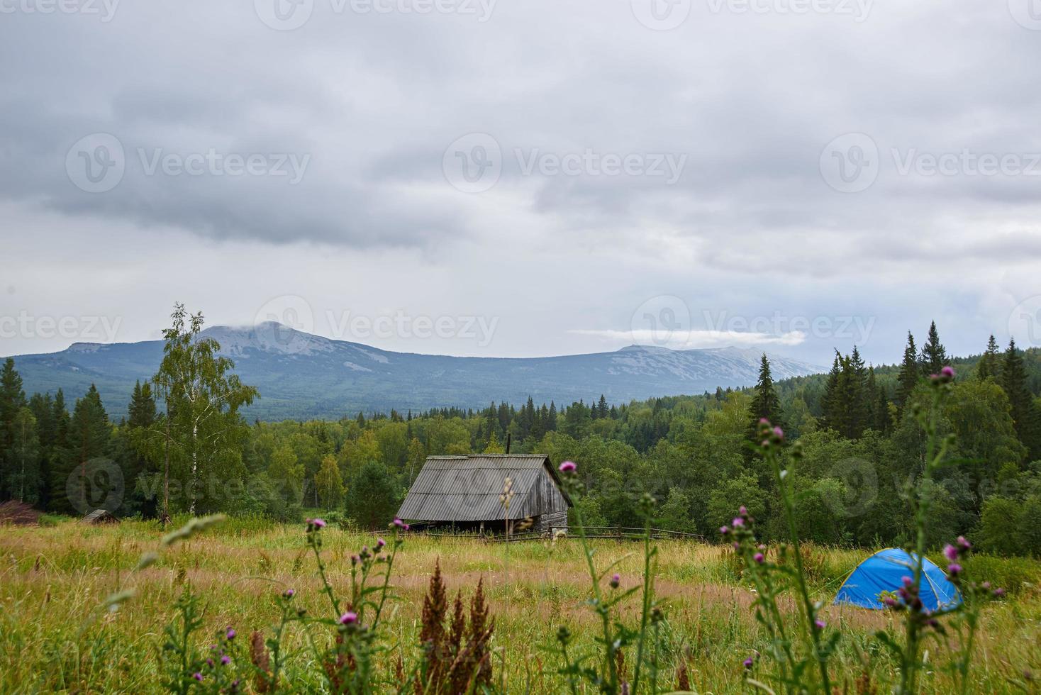 paisagem, prado verde, tenda azul, casa de madeira, gramíneas de campo, bela vista dos picos das montanhas nas nuvens. foto