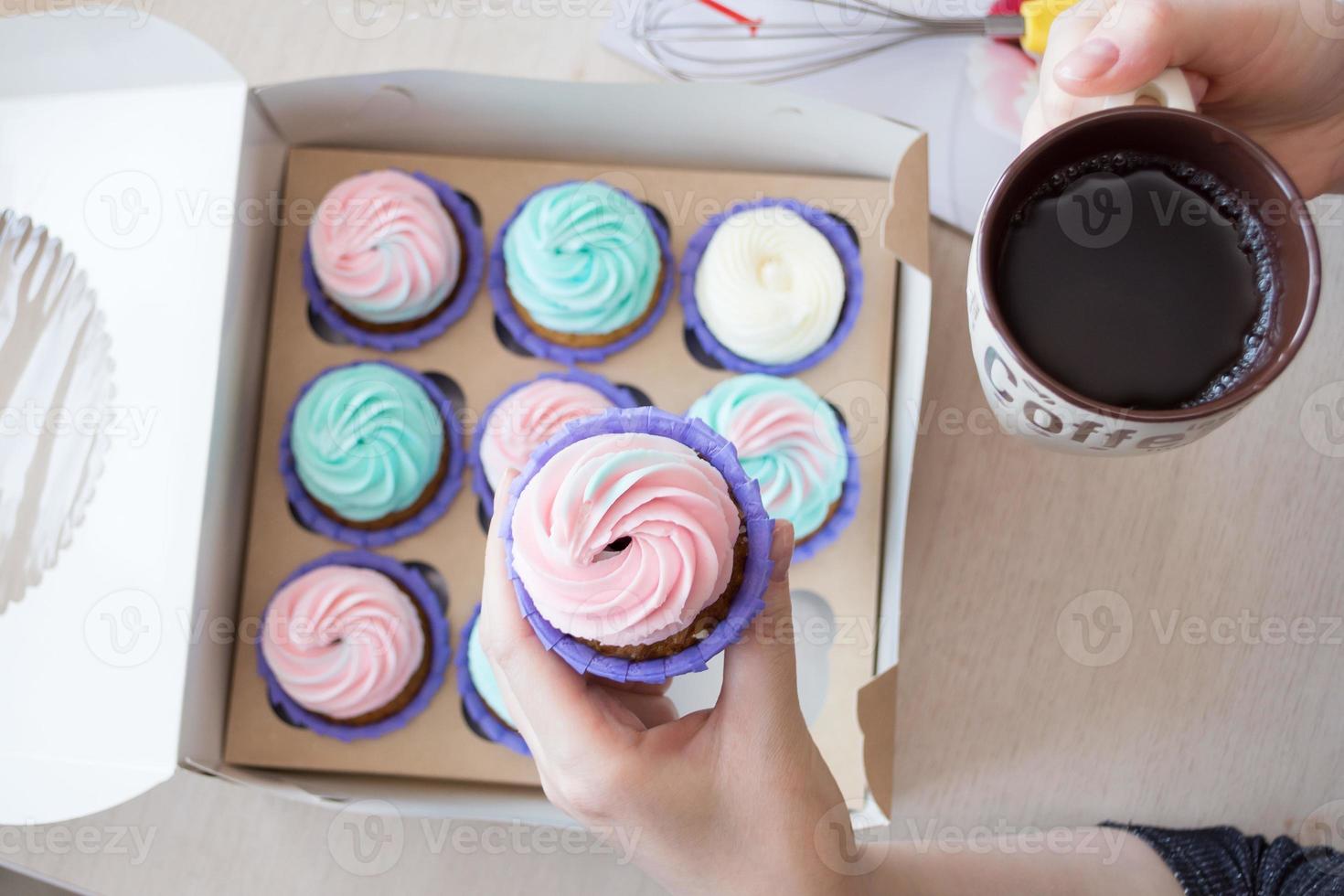 cupcake com creme rosa e azul nas mãos de uma garota no fundo de uma caixa de cupcakes e café foto