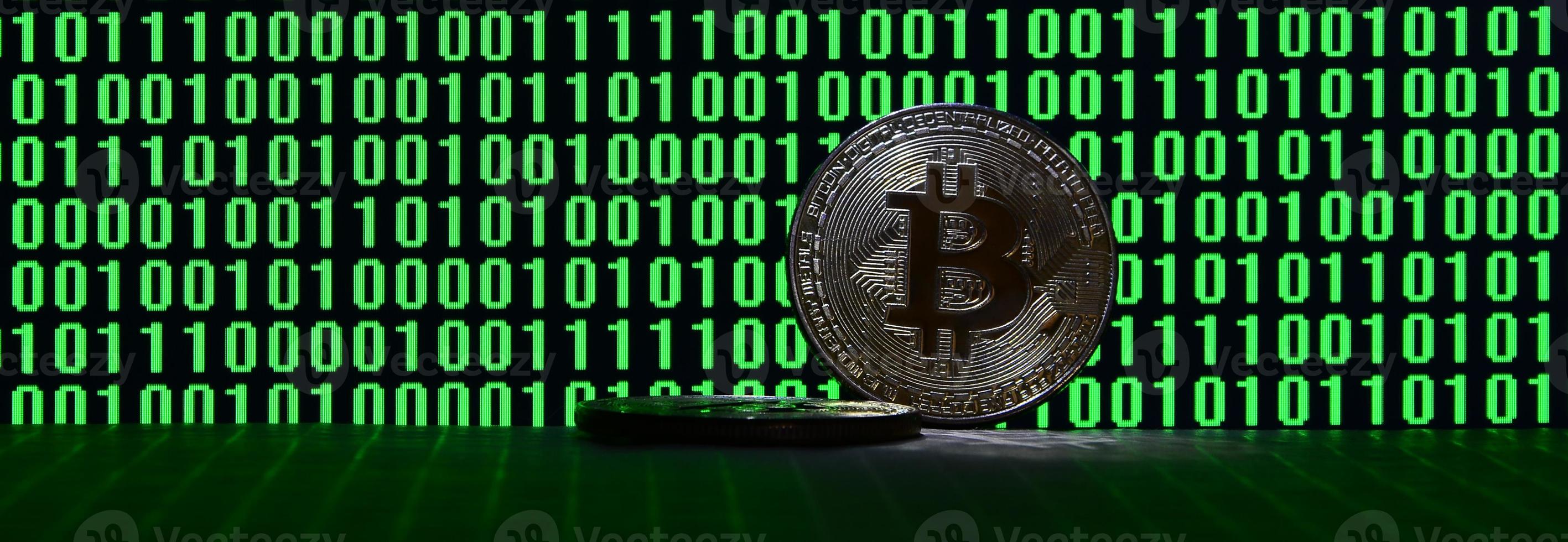 um par de bitcoins está em uma superfície de papelão no fundo de um monitor representando um código binário de zeros verdes brilhantes e unidades de um em um fundo preto. iluminação baixa chave foto