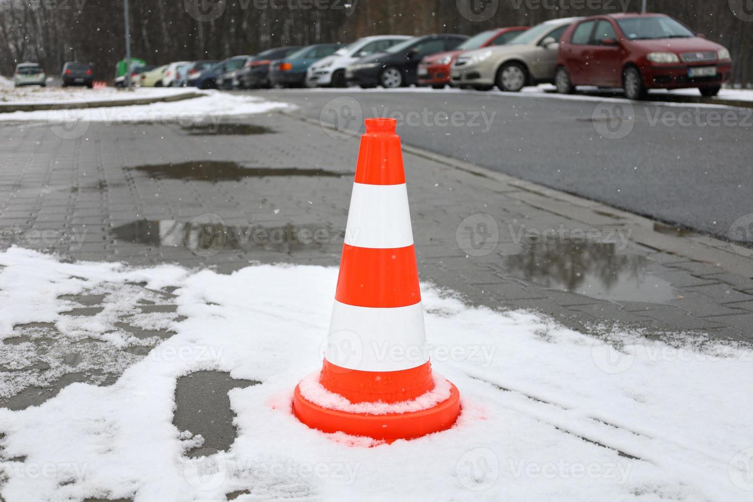 cones de trânsito laranja brilhante em pé na linha lateral, coberto de neve branca fresca. início da queda de neve, flocos de neve desfocados em primeiro plano. carros estacionados no fundo, irreconhecíveis. foto