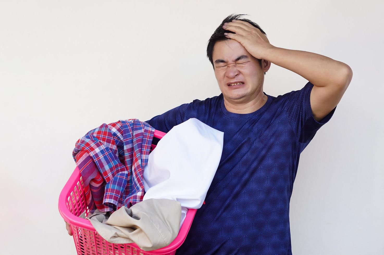 homem asiático segura cesta de pano para lavanderia, sente-se preguiçoso, irritado, cansado, coloca a mão na cabeça. conceito, tarefa diária, trabalho doméstico. homem não quer lavar roupa. fazer cara engraçada. foto