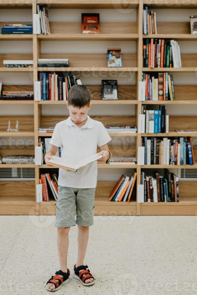 um menino está na biblioteca e lê um livro em pé. preparando-se para a lição de casa. o menino adora ler. espaço livre na escola. aprendizagem extracurricular. foto