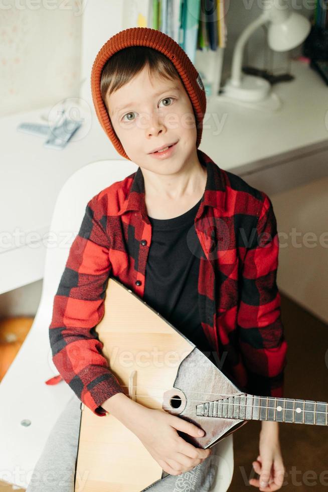 menino de chapéu vermelho e camisa xadrez toca balalaica. menino bonito segurando seu violão. aulas de música em casa. passatempo para a alma. ensino de musica em casa foto