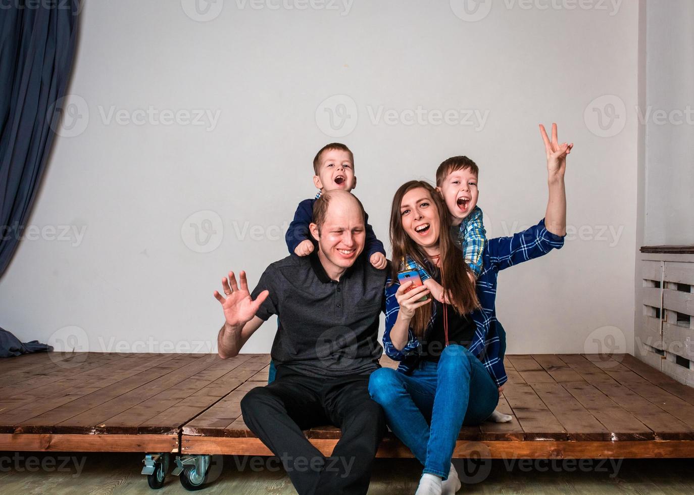 família sorridente segurando a forma da casa enquanto está sentado no chão de madeira em casa. photoshoot de uma família com 2 filhos, fundo branco. relações familiares calorosas. pais jovens foto