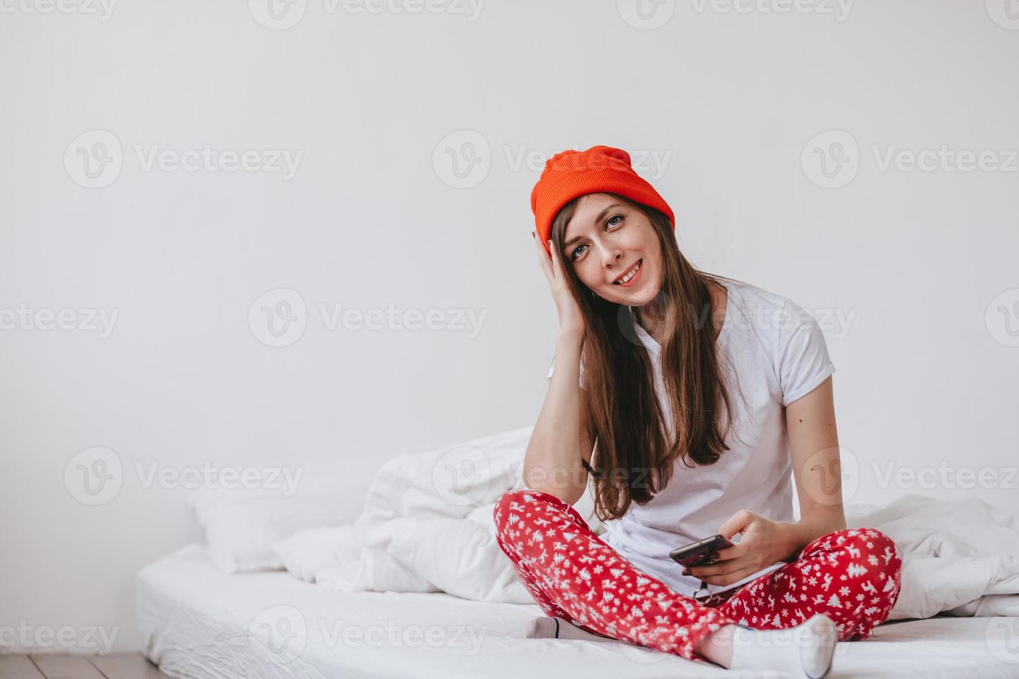 garota engraçada sorridente em um chapéu vermelho e pijama de natal, sentado na cama com lençóis brancos. feriados para o natal. garota de bom humor natalino. garota em uma camiseta branca e pijama foto