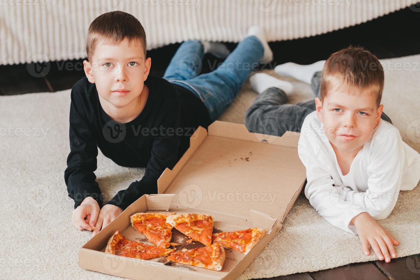 dois meninos estão deitados no chão comendo pizza de calabresa de uma caixa. quadro vertical. dois meninos estão deitados no chão comendo pizza de calabresa de uma caixa. foto