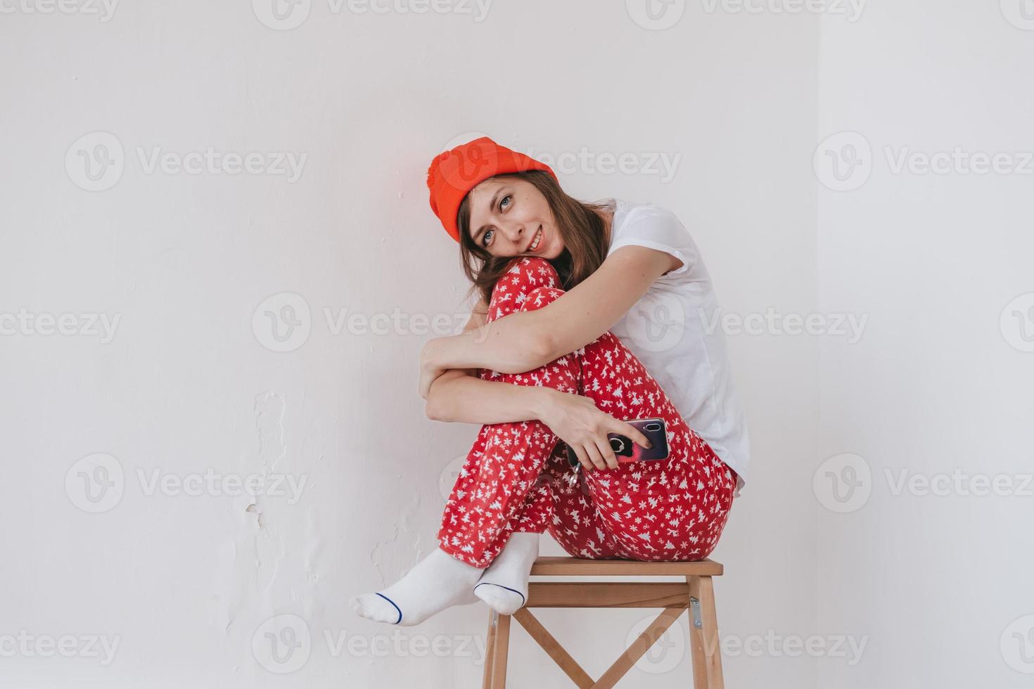 garota engraçada sorridente em um chapéu vermelho e pijama de natal, sentado na cama com lençóis brancos. feriados para o natal. garota de bom humor natalino. garota em uma camiseta branca e pijama foto