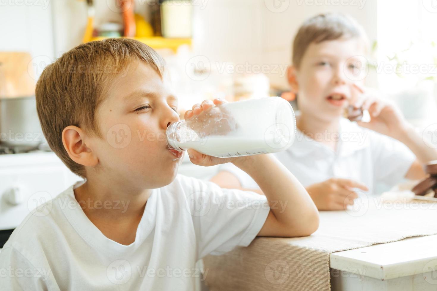 um menino está bebendo leite de uma garrafa na cozinha de casa. café da manhã com leite. criança feliz em uma camiseta branca bebe leite de uma garrafa foto