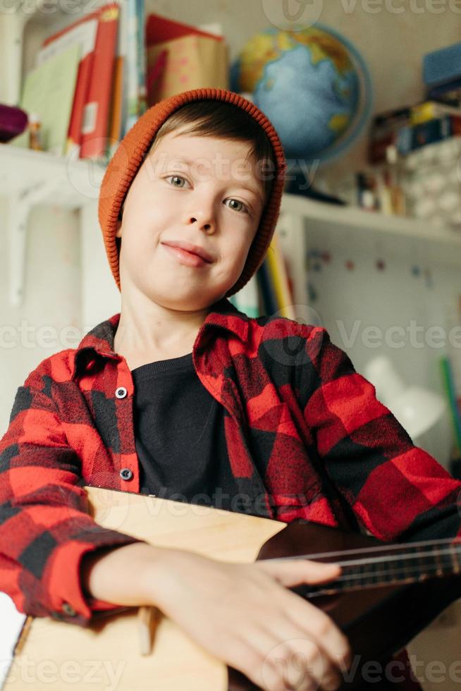 menino de chapéu vermelho e camisa xadrez toca balalaica. menino bonito segurando seu violão. aulas de música em casa. passatempo para a alma. ensino de musica em casa foto