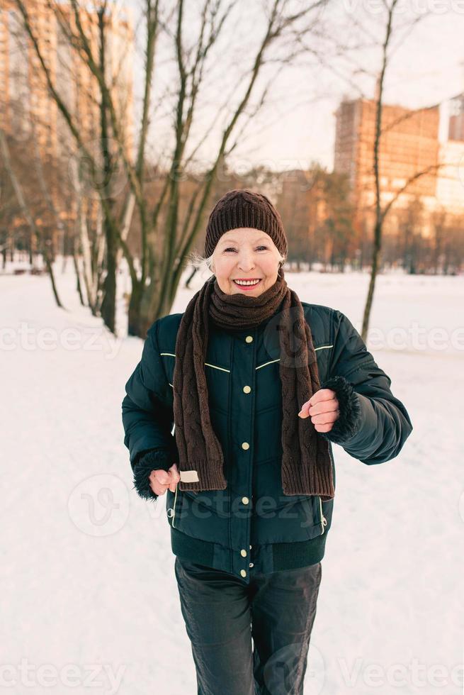 mulher sênior de chapéu e jaqueta esportiva correndo e fazendo exercícios esportivos no parque de inverno de neve. inverno, idade, esporte, atividade, conceito de temporada foto