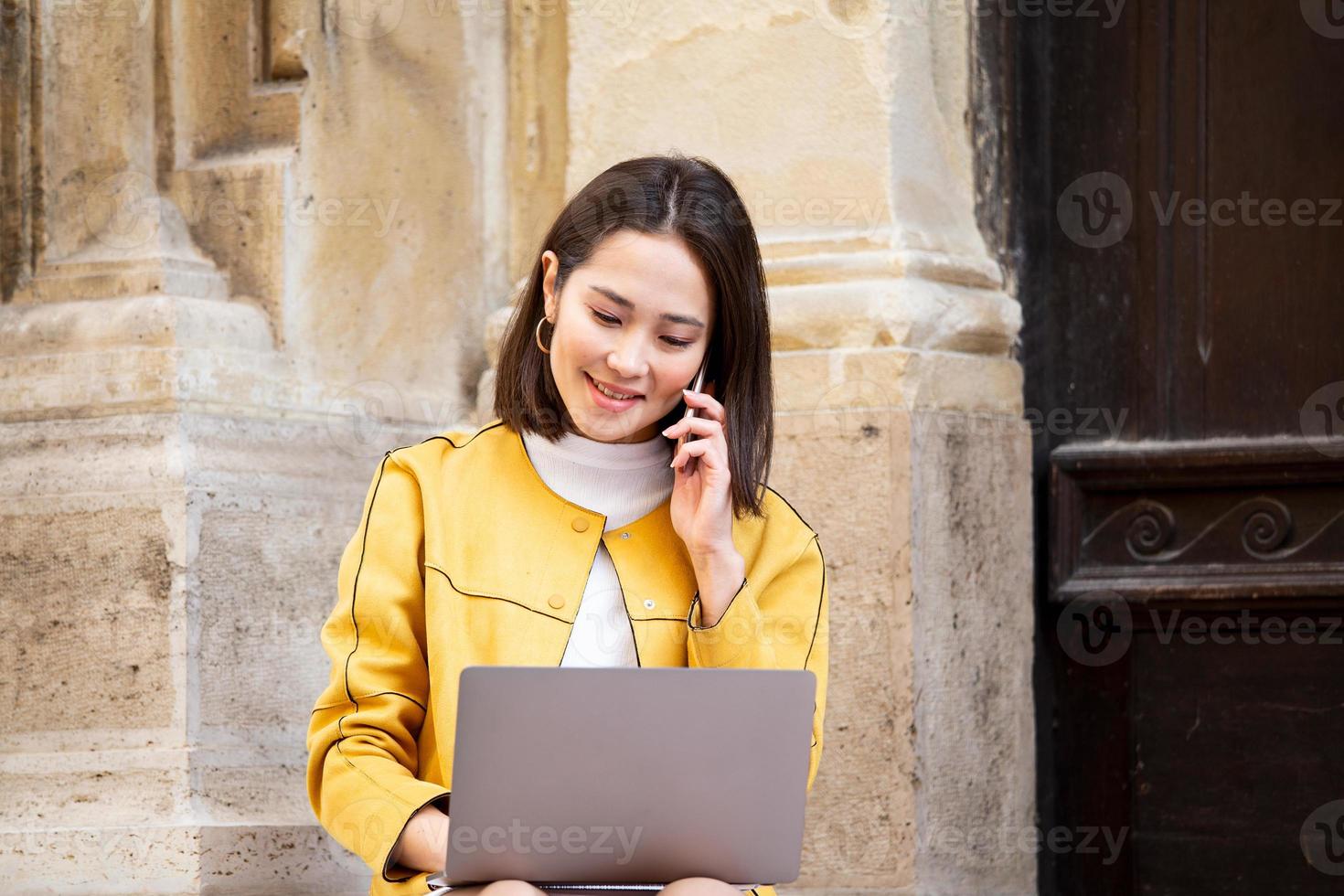mulher asiática sorridente trabalhando no computador portátil enquanto está sentado no chão e falando em seu telefone inteligente. mulher asiática falando no celular móvel enquanto estiver usando computador portátil foto