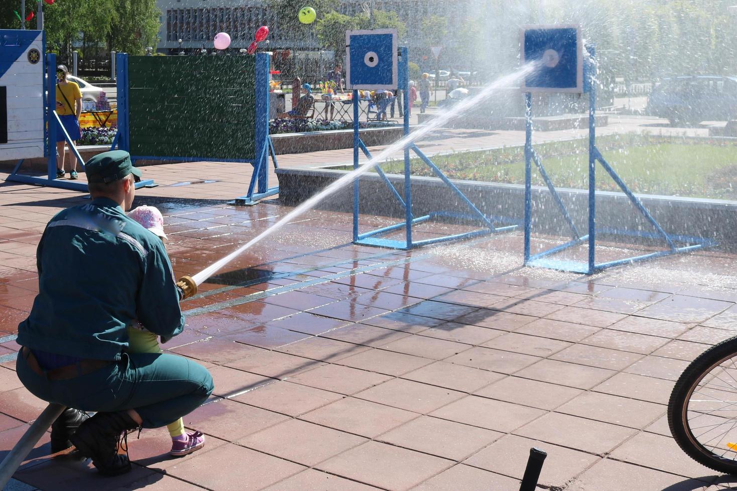 o homem de um bombeiro está ensinando uma criança, uma menina a apagar um incêndio atirando um jato d'água de um bronzboyt, uma mangueira de incêndio, uma mangueira para um alvo foto