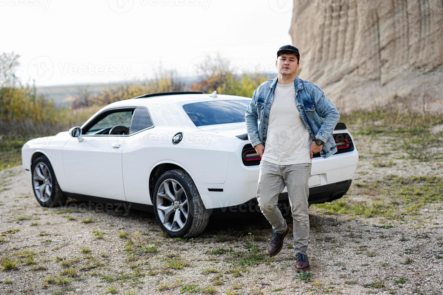 homem bonito em jaqueta jeans e boné andando perto de seu muscle car branco na carreira. foto