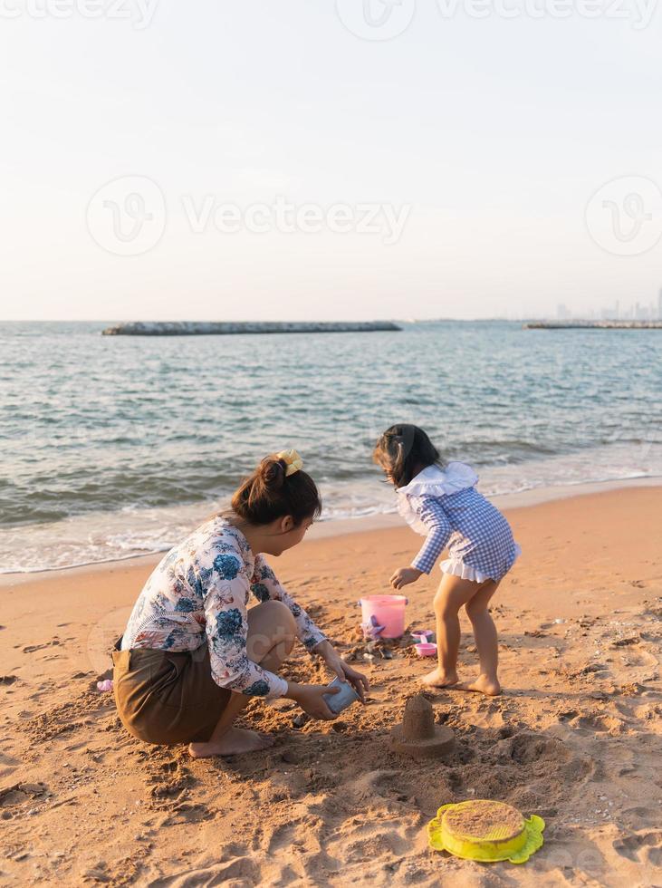 menina bonitinha asiática e sua mãe brincando ou fazendo castelo de areia ou cavando com areia na praia tropical. crianças com lindo mar, céu azul de areia. crianças felizes em férias à beira-mar na praia. foto