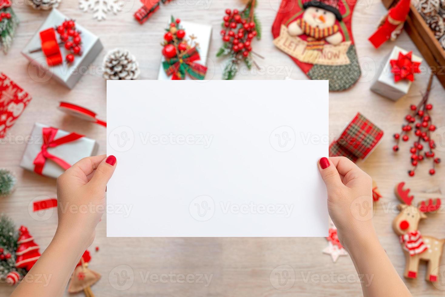 mãos de mulher segurando uma folha de papel em branco para adicionar o texto do cartão. cena de natal com muitos presentes e decorações na mesa de madeira foto