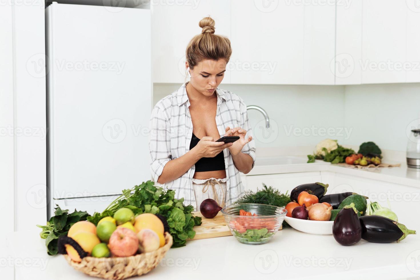 mulher com fones de ouvido sem fio está usando smartphone durante o cozimento na cozinha branca moderna foto