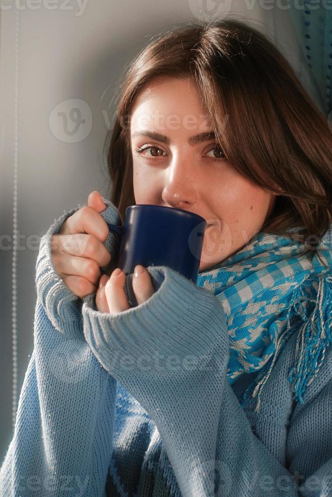 mulher bonita e triste ficando em casa por causa da gripe e temperatura no suéter quente e cachecol com xícara de chá perto da janela foto