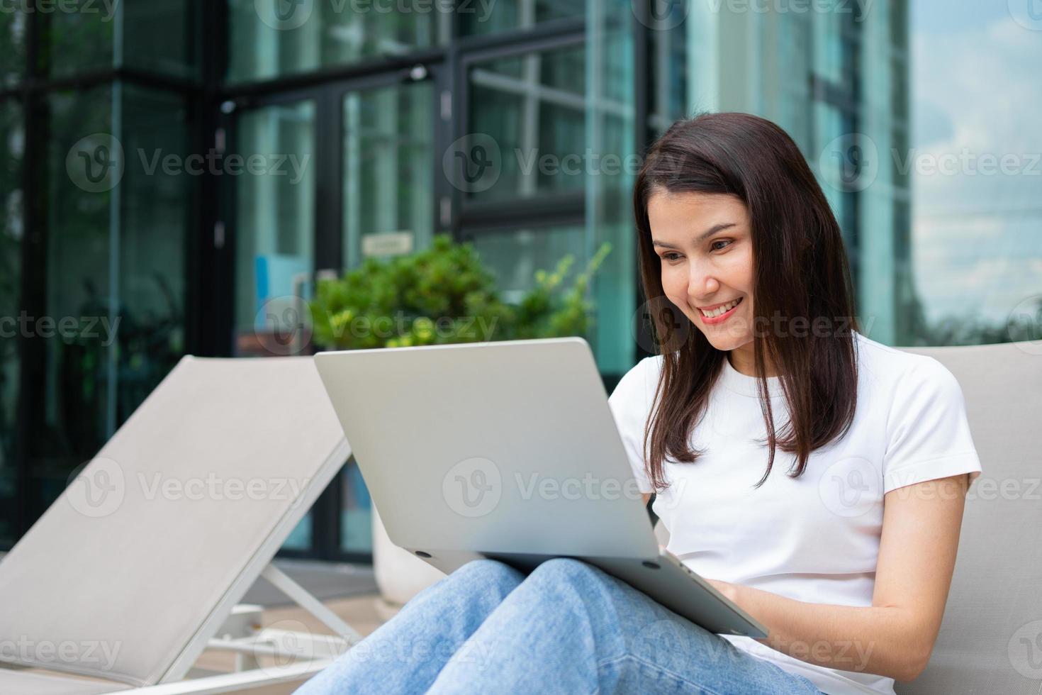 mulher jovem empreendedora feliz sentada na cama de bronzeamento ao lado da piscina e usando o laptop para trabalhar on-line remoto, projeto de negócios on-line no quintal tranquilo da casa de resort, trabalhar de férias foto