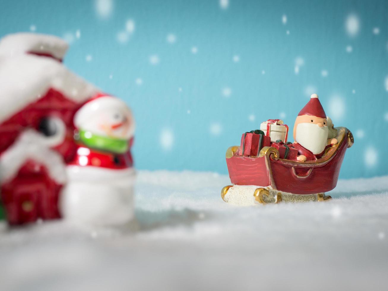 feliz papai noel com caixa de presentes no trenó de neve indo para a casa de neve. perto da casa de neve tem boneco de neve e árvore de natal. papai noel e casa de neve na neve o fundo é azul em pó. foto