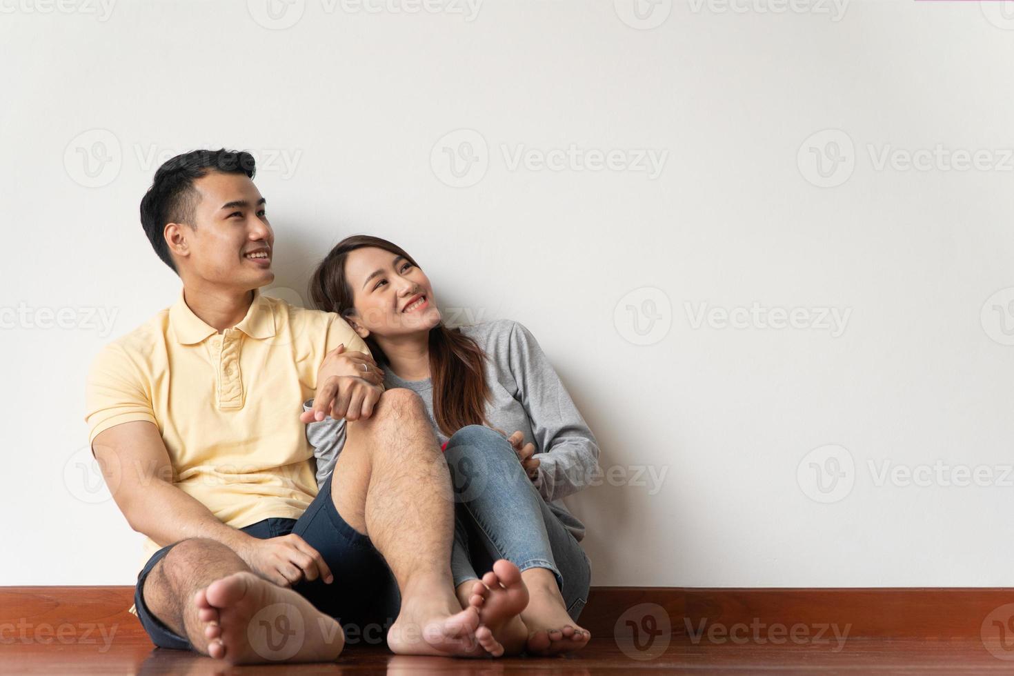 casal jovem asiático feliz sentado no chão em casa e olhando para cima com espaço de cópia apontando para o ar no fundo da parede branca foto