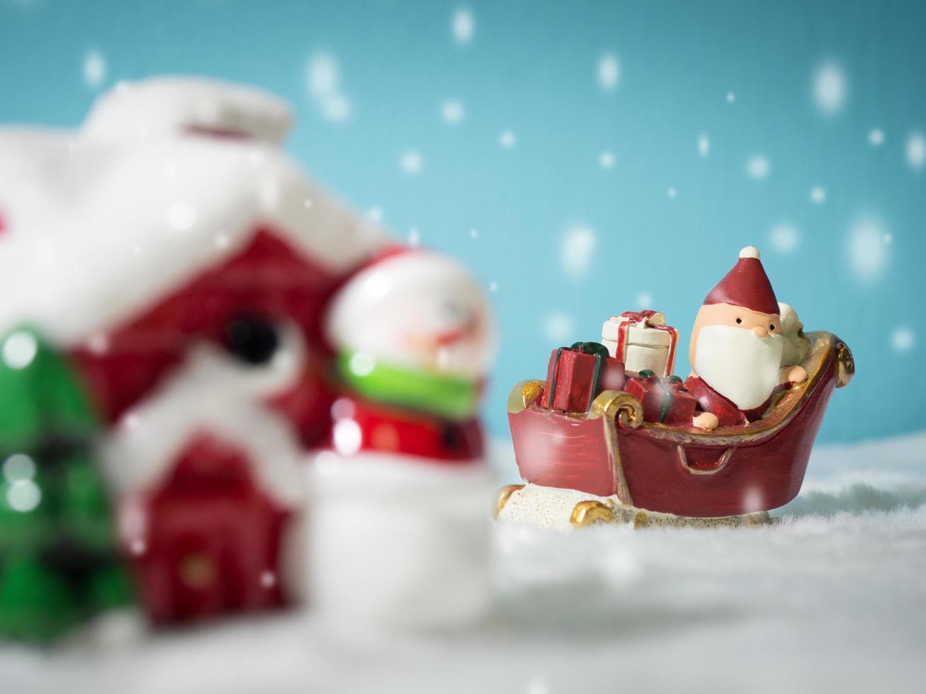 feliz papai noel com caixa de presentes no trenó de neve indo para a casa de neve. perto da casa de neve tem boneco de neve e árvore de natal. papai noel e casa de neve na neve o fundo é azul em pó. foto