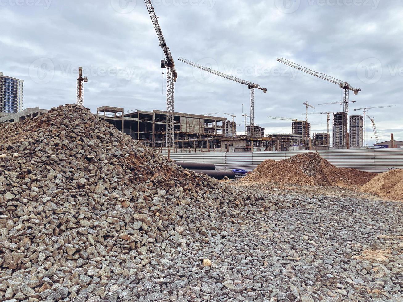 construção de casas altas na cidade. guindastes puxam lajes de concreto pesadas para a construção de edifícios residenciais. pedras para estradas são derramadas ao redor foto