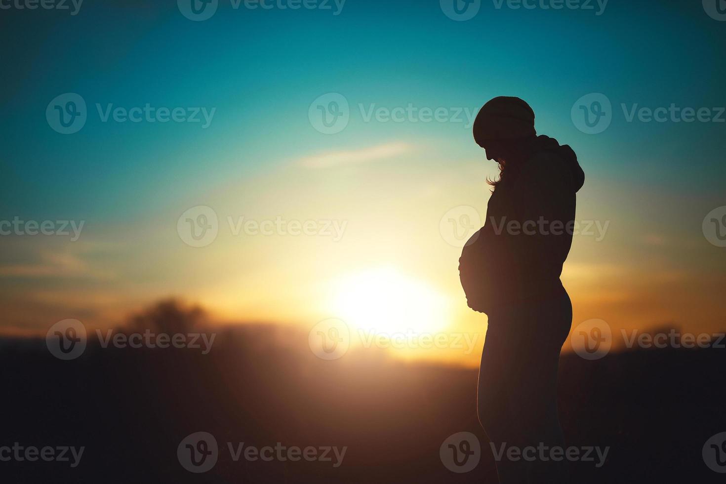 silhueta de uma futura mãe, mulher grávida relaxa no parque em um fundo por do sol foto