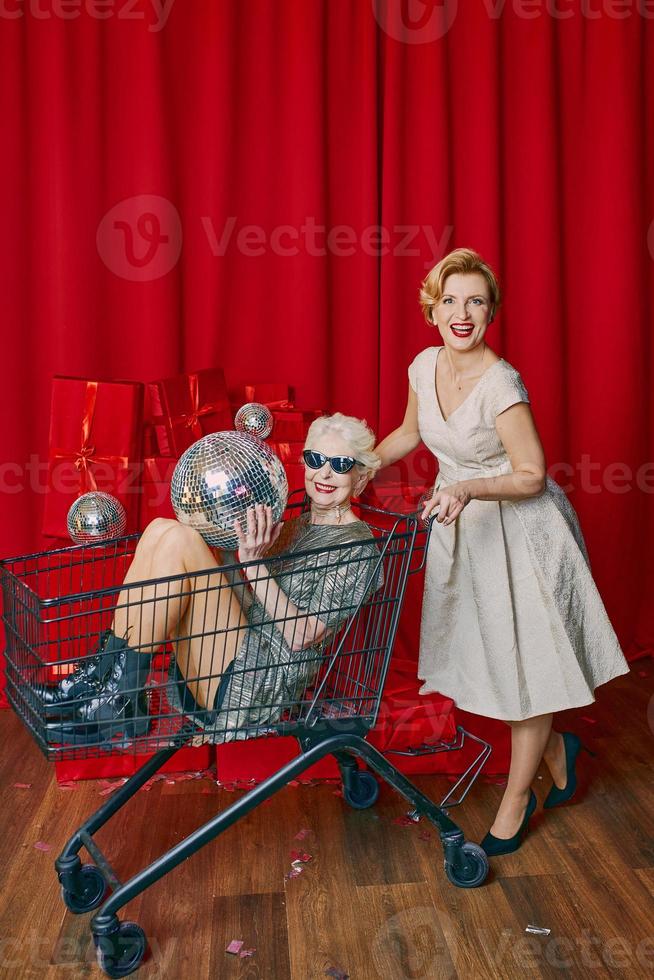 mulher madura rolando elegante mulher sênior em óculos de sol e prata veste o carrinho de supermercado na festa. festa, discoteca, celebração, conceito de terceira idade foto