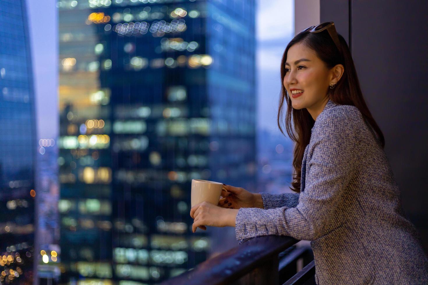 jovem asiática apreciando a vista noturna de sua varanda enquanto olha para a paisagem urbana de arranha-céus à noite com uma xícara de café quente para habitação e conceito de estilo de vida moderno foto
