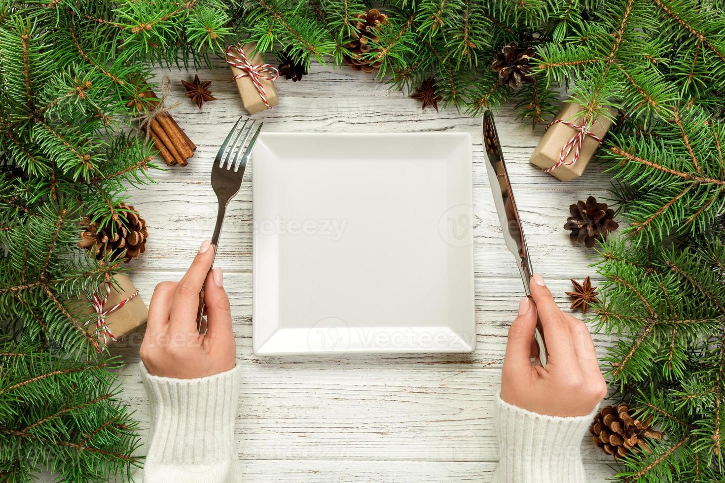 garota de vista superior mantém garfo e faca na mão e está pronta para comer. prato quadrado branco vazio sobre fundo de madeira de natal. conceito de prato de jantar de férias com decoração de ano novo foto