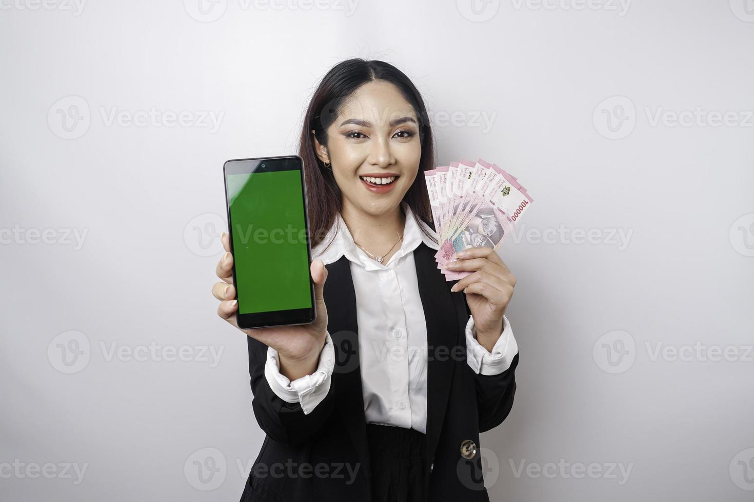 uma jovem empresária feliz está vestindo terno preto, mostrando seu telefone e dinheiro em rupia indonésia isolado por fundo branco foto