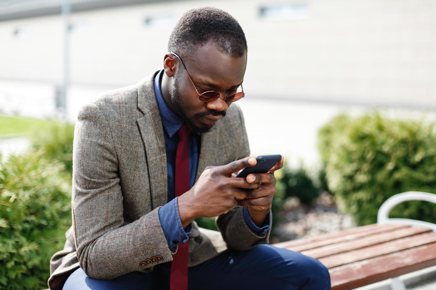 homem de negócios afro-americano trabalha em seu smartphone sentado no banco do lado de fora foto