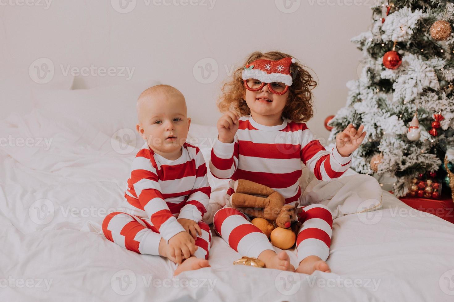 crianças de pijama vermelho e branco experimentam óculos engraçados com papai noel sentado na cama. estilo de vida. irmão e irmã comemorando o natal. menino e menina estão brincando em casa. foto de alta qualidade