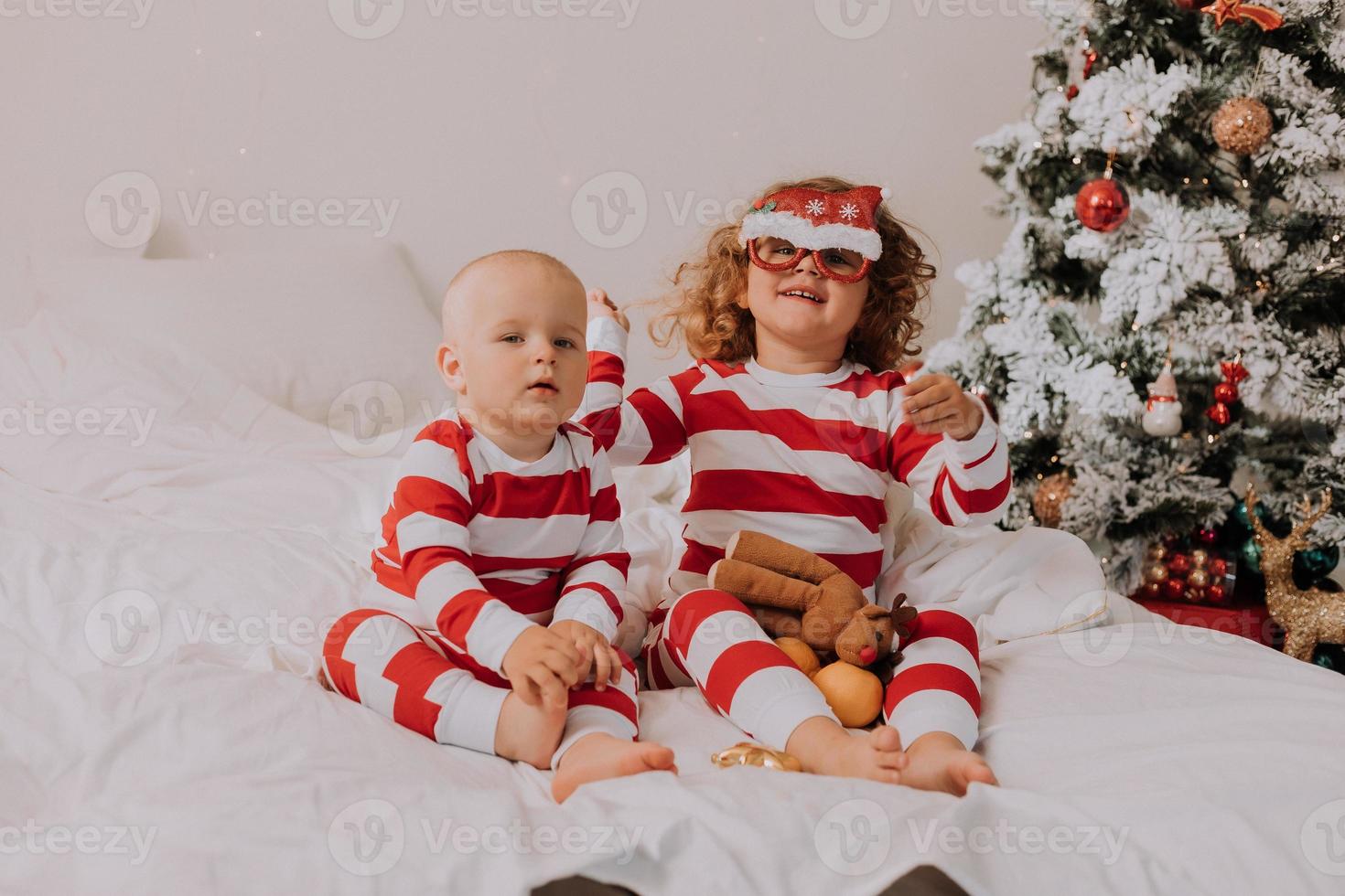 crianças de pijama vermelho e branco experimentam óculos engraçados com papai noel sentado na cama. estilo de vida. irmão e irmã comemorando o natal. menino e menina estão brincando em casa. foto de alta qualidade